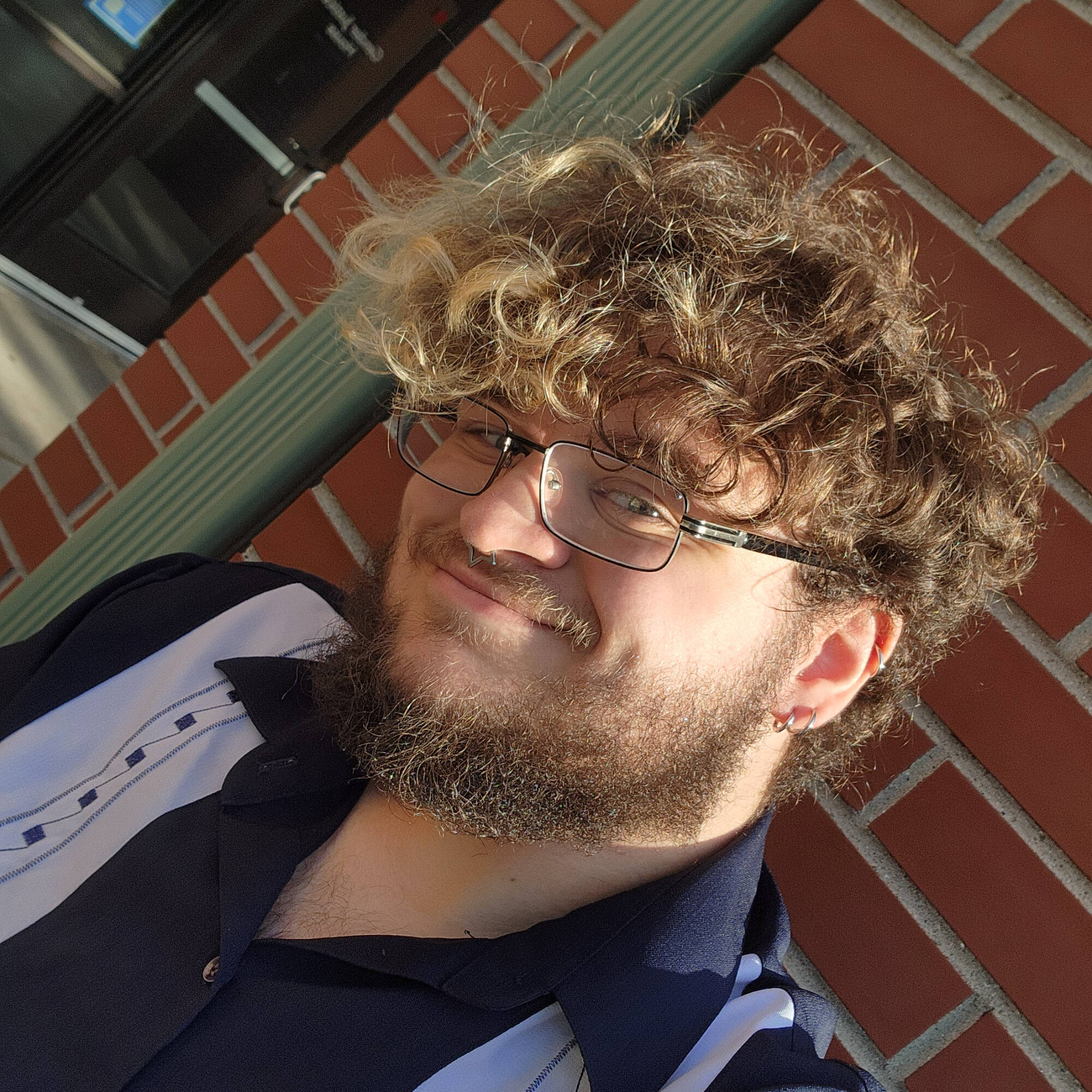 Marshall has curly brown hair and a beard, with glasses. He is smiling with the sunlight falling on his face and a brick background.